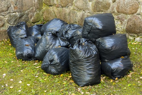 A pile of ten black rubbish bags made of glossy plastic, tightly sealed, and stacked on a grassy area in front of a rough stone wall. The bags contain garden waste or refuse, with some bags slightly bulging from their contents. The grass under the bags is green with scattered small leaves and pebbles, indicating an outdoor setting likely associated with garden clearance. The stone wall behind the bags is composed of irregularly shaped rocks with moss growth, providing a textured backdrop. The scene appears to be part of a yard or garden space that requires waste disposal services, typical of garden clearance jobs in Greenwich. The overall lighting suggests natural daylight, making the details and textures clearly visible, aligning with rubbish removal activities handled by Garden Clearance Greenwich.