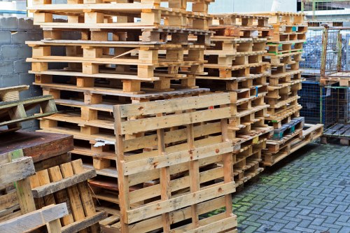 A large stack of unfinished wooden pallets is neatly arranged outdoors on a paved surface in a storage or industrial area, with some pallets leaning against each other and other smaller pallets and crates positioned nearby. The pallets are made of light-colored, untreated timber with visible grain and rough textures, showing gaps between the slats and some uneven edges. The setting includes a brick wall and a metal fence in the background, indicating a typical site for rubbish or waste collection related to construction or commercial activity. The overall scene conveys a clean, organized arrangement suitable for rubbish removal services, such as those offered by Garden Clearance Greenwich, in the Greenwich area of London, with a focus on timber waste management.