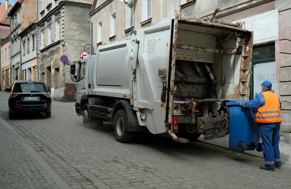 A middle-aged man with dark hair, wearing a black T-shirt with white text on the front, is seen bending over to dispose of waste into a stainless steel public rubbish bin situated on a paved sidewalk.
