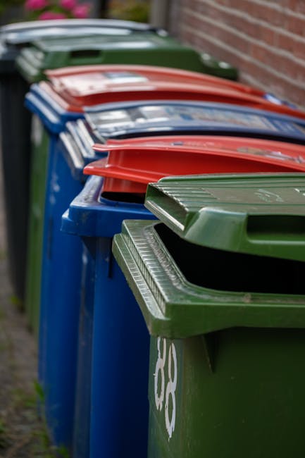 A row of large outdoor wheelie bins lined up along a pavement adjacent to a brick wall, featuring predominantly blue bins with green, red, and blue lids. The bins are made of sturdy plastic with textu