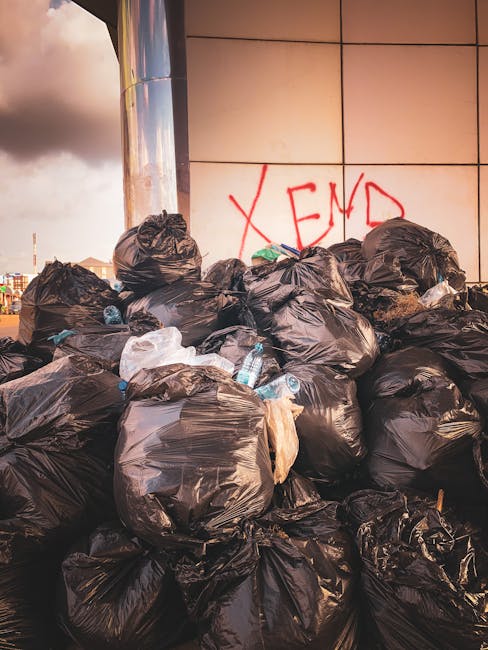 A row of large outdoor wheelie bins lined up along a pavement adjacent to a brick wall, featuring predominantly blue bins with green, red, and blue lids. The bins are made of sturdy plastic with textu